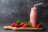 A glass bottle of strawberry smoothie with a pink straw, surrounded by fresh strawberries and mint leaves on a wooden cutting board.